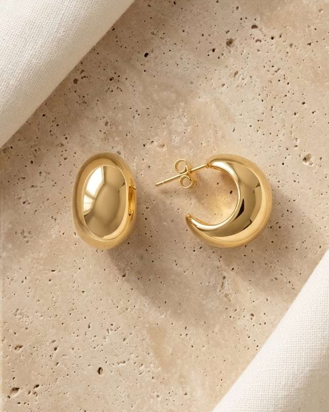 A pair of polished gold dome huggie earrings displayed on a beige stone slab with draped cream fabric around it.