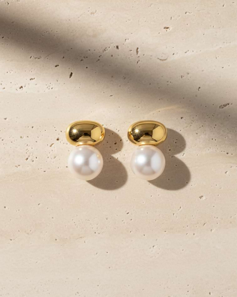 A pair of gold stud earrings with white pearl drops, displayed on a textured beige stone surface with soft diagonal shadows.
