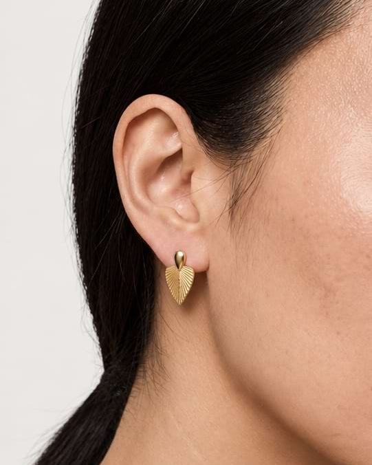 A close-up side profile of a woman wearing a small gold leaf stud earring on her earlobe against a light neutral background.