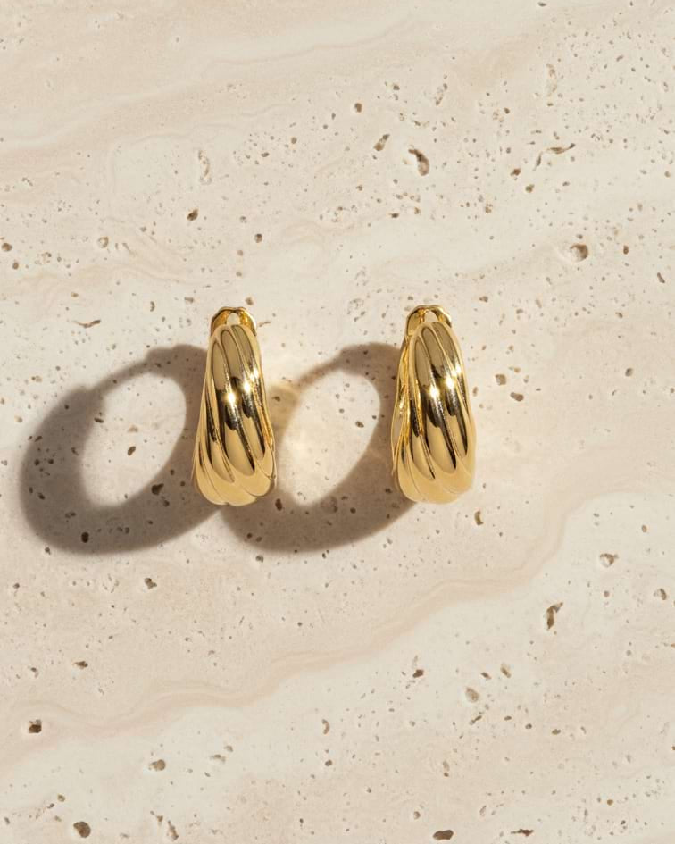 A pair of gold hoop earrings with a textured, ribbed surface, displayed on a light beige stone background with soft shadows.