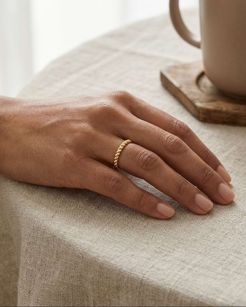 A close-up of a hand resting on a beige linen tablecloth, wearing a delicate gold ring with small round beads. A matching beige ceramic mug sits on a wooden coaster in the background.