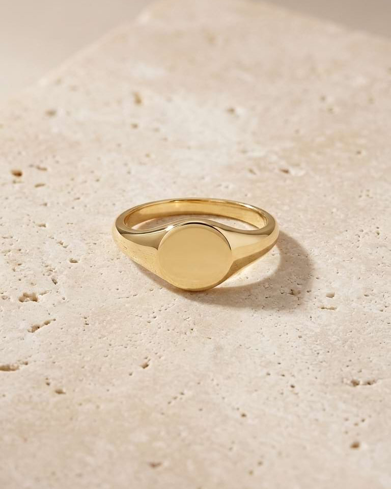 A polished yellow gold signet ring with a flat oval face, resting on a textured beige stone surface under warm, directional sunlight.