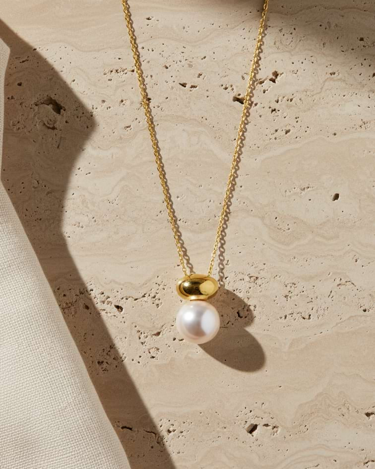 A gold chain necklace with a pendant featuring a white pearl and a polished gold bead, displayed on a textured beige stone surface with soft shadows.
