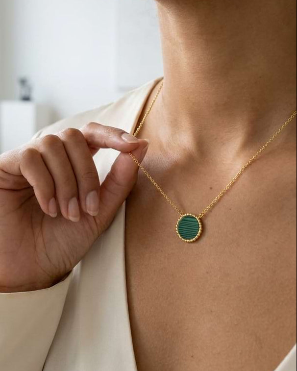 A close-up lifestyle shot of a woman wearing a delicate gold chain necklace with a round green malachite pendant, styled with a cream-colored blouse in a bright, modern room.