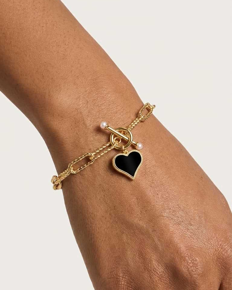 A gold chain bracelet with a black heart charm and small pearl-like beads, shown worn on a person's wrist against a light neutral background.