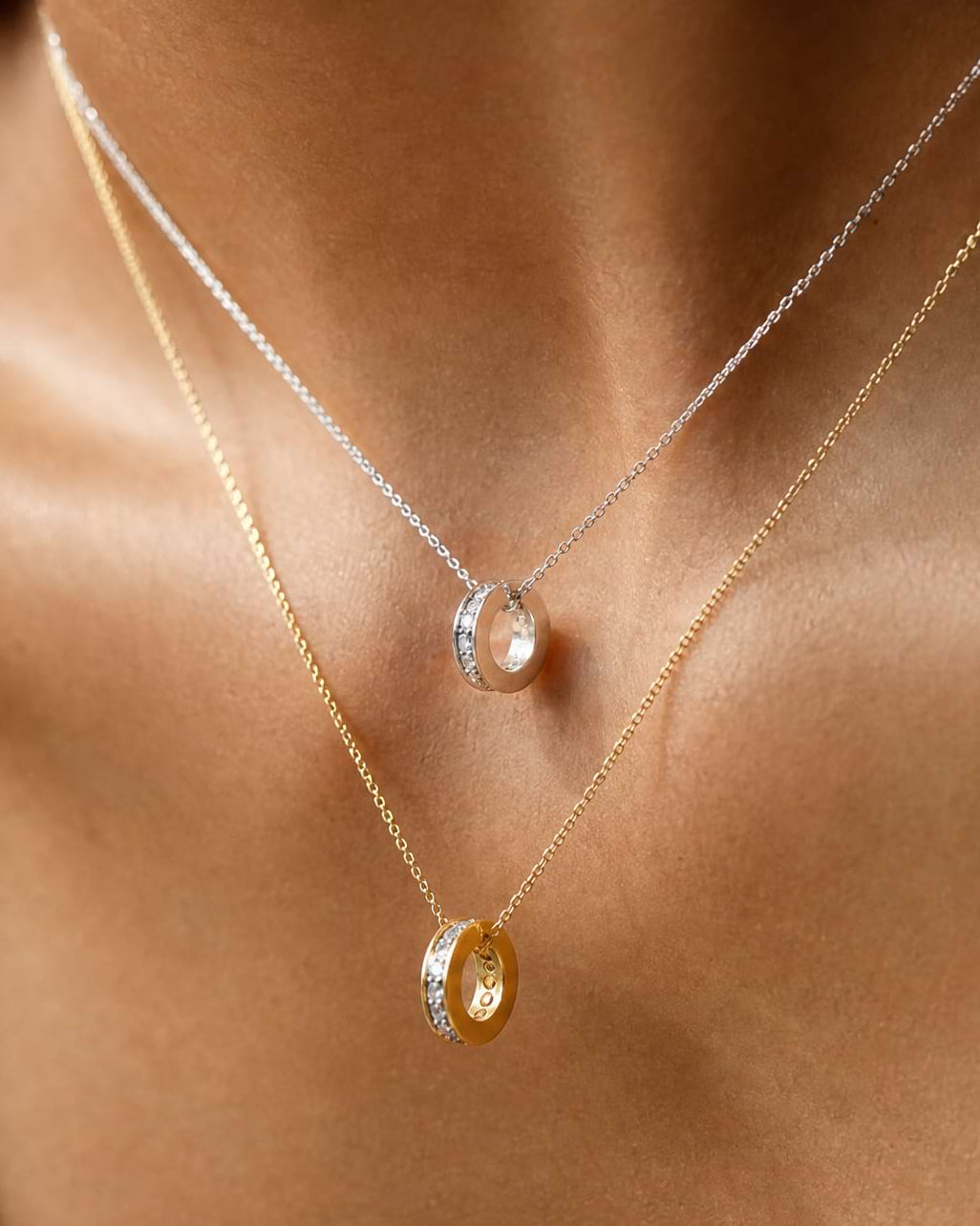 A close-up of a model's neck and chest wearing two delicate layered necklaces. The top necklace is silver with a small oval clear stone pendant, and the bottom necklace is gold with a larger gold oval pendant featuring a small clear accent.