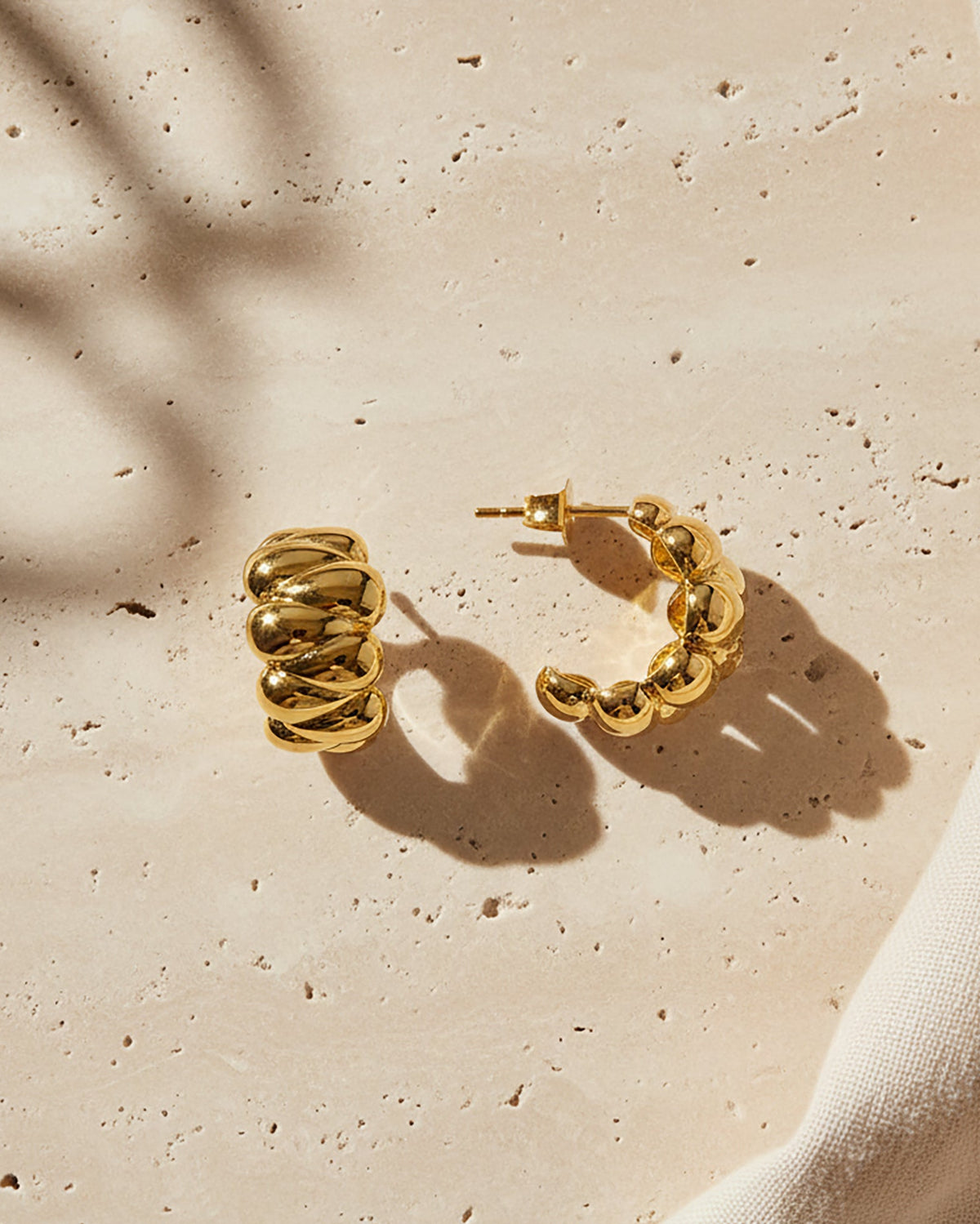 A pair of gold puffy huggie earrings with a textured, ridged spiral design, displayed on a beige stone surface with soft leaf shadows.
