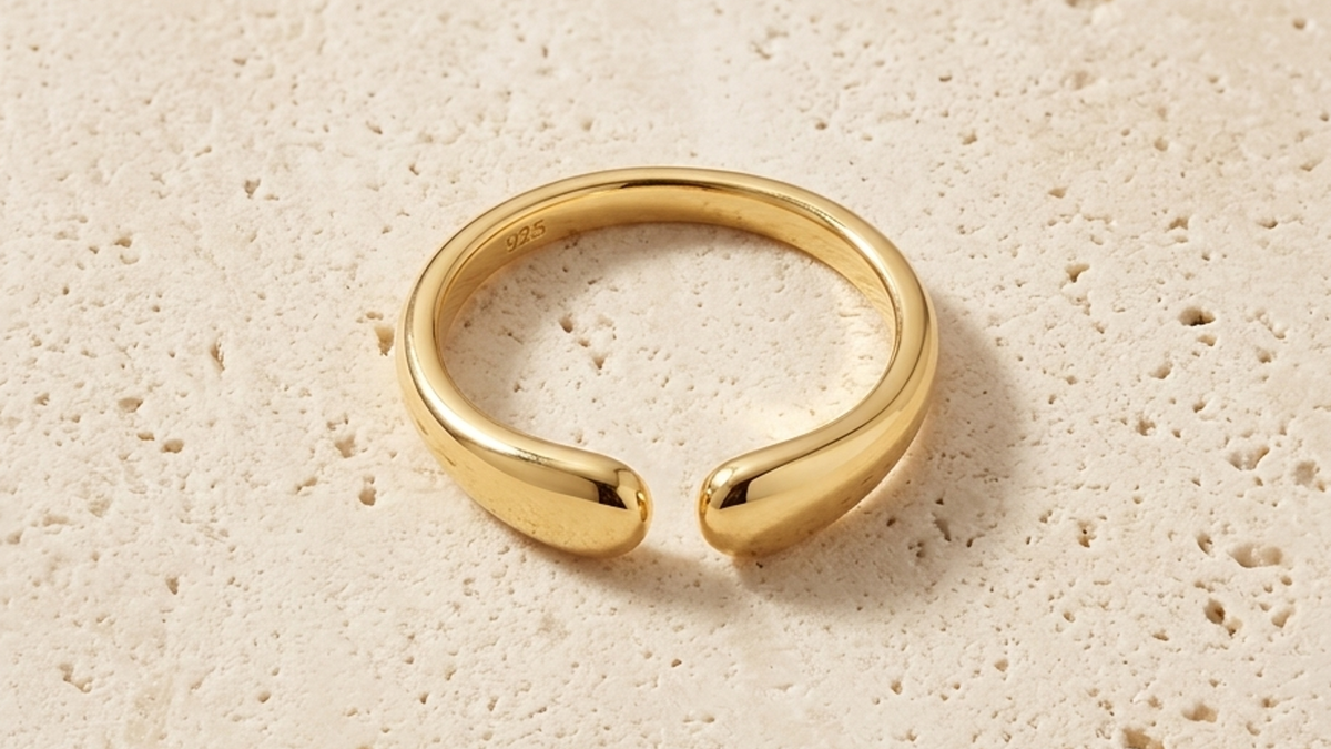 A polished gold open ring with a smooth, organic curved shape, resting on a textured beige stone surface.