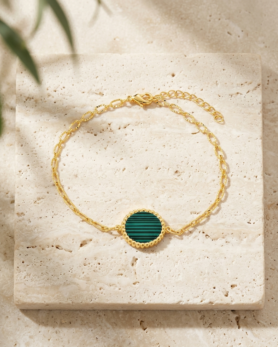 A delicate gold chain bracelet with a round malachite stone inlay, displayed on a textured beige stone slab with soft leaf shadows.