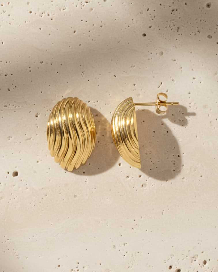 A pair of gold crescent-shaped hoop earrings with a textured, ridged surface, displayed on a light beige textured stone background with soft shadows.