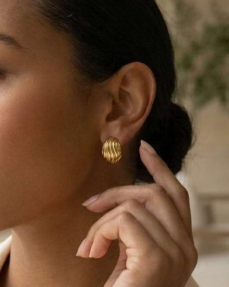 A woman in a cream satin shirt wearing a textured gold stud earring, shown in a profile view in a softly lit indoor setting.