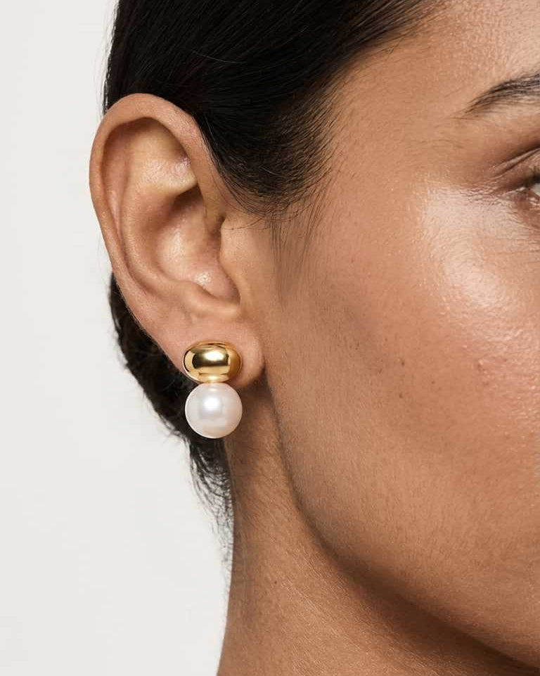 A close-up of a model's ear wearing a gold and white pearl stud earring against a light neutral background.