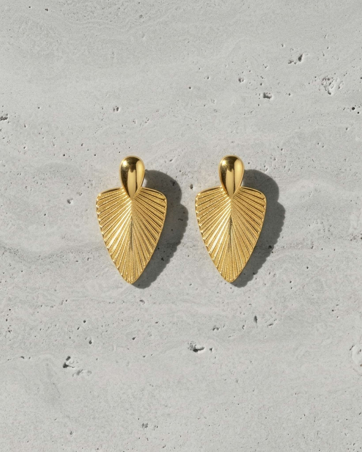 A pair of textured gold leaf-shaped drop earrings displayed on a light gray concrete surface.