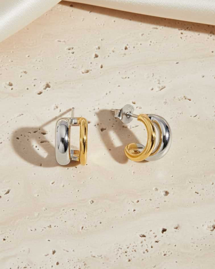 A pair of double-hoop earrings in polished white and yellow gold, displayed on a textured beige stone surface with a draped cream satin fabric in the background.