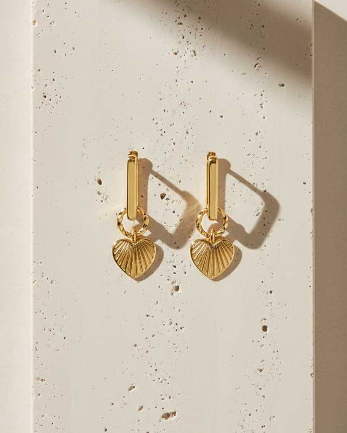 Gold earrings with a dangling heart on a textured white surface