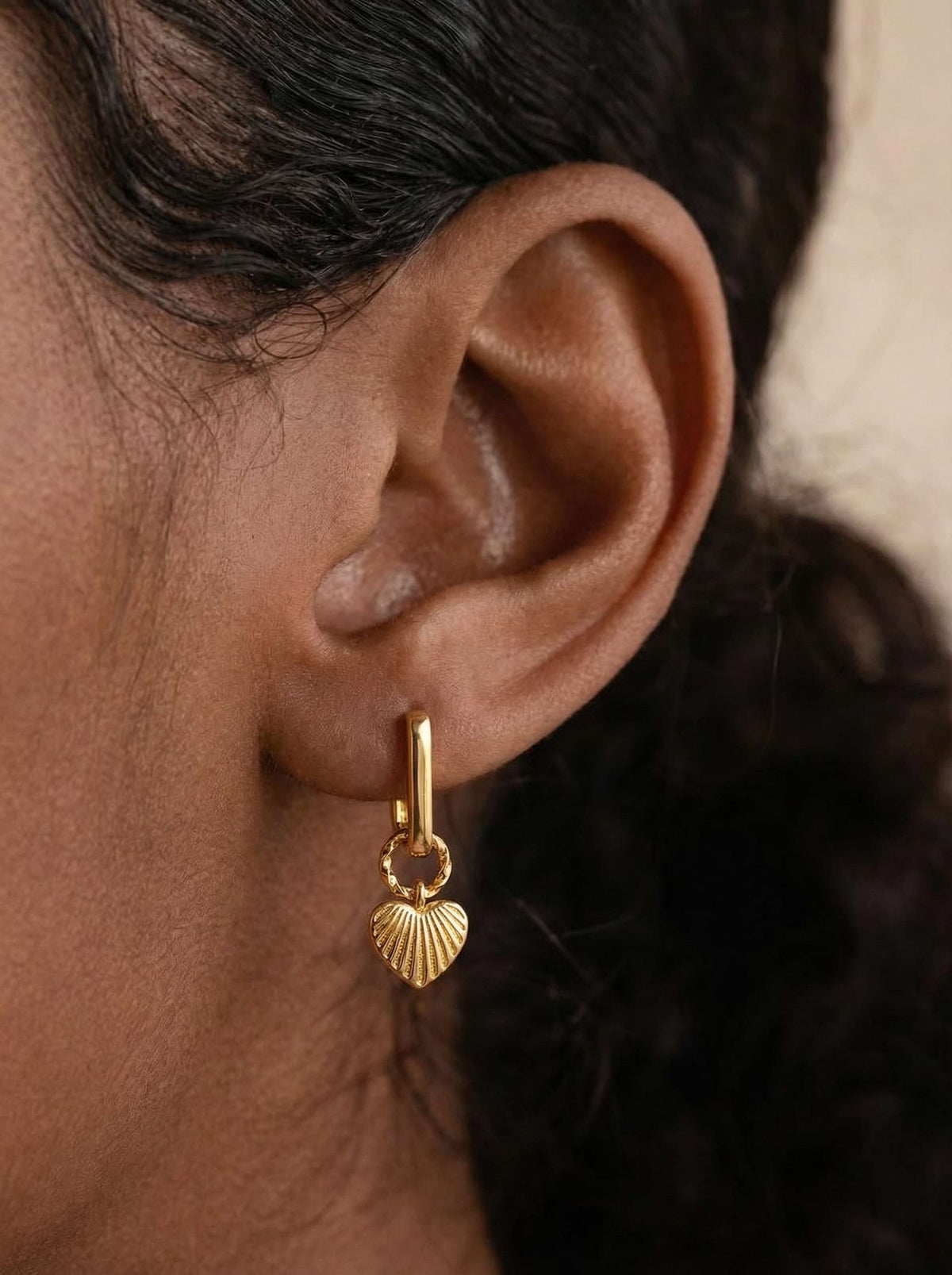 A close-up of a woman's ear wearing a small gold huggie hoop earring with a dangling ribbed gold heart charm.