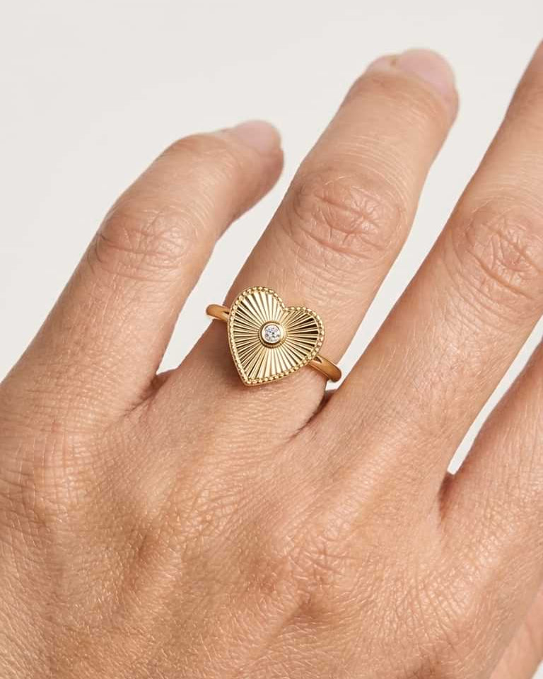 A gold ring with a textured, sunburst heart-shaped face and a small central clear stone, shown worn on a person's finger against a light neutral background.
