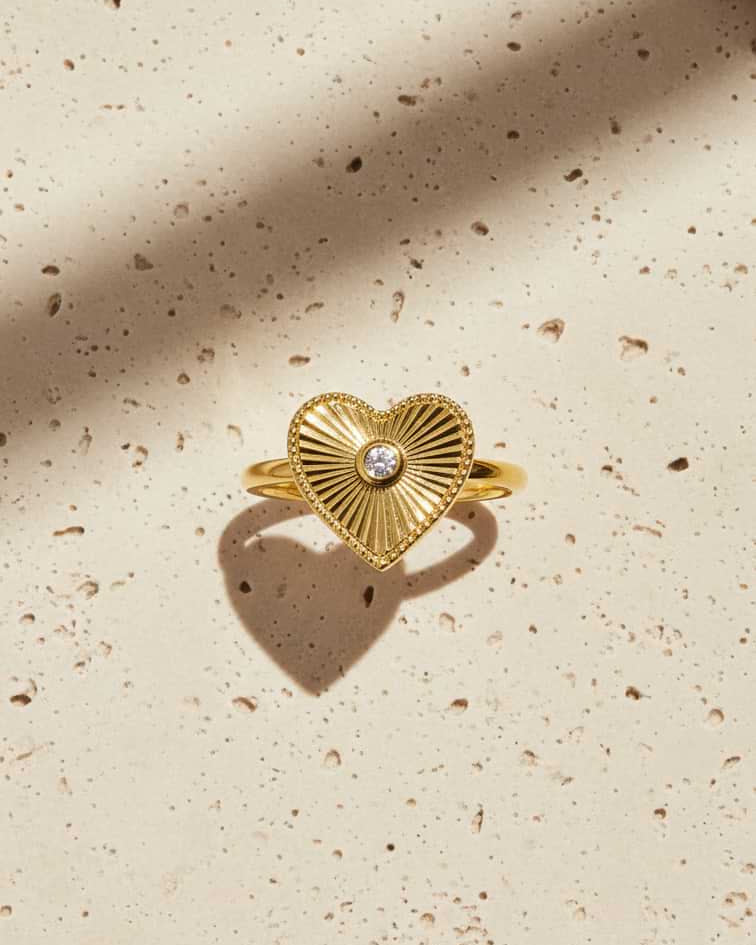 A gold heart-shaped ring with a sunburst textured pattern and a small central clear gemstone, displayed on a speckled beige surface with soft shadows.
