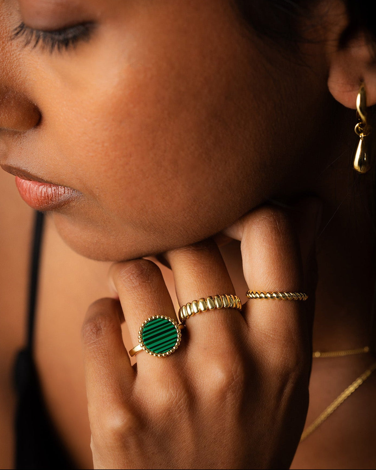 Close-up of a person wearing gold rings, including a prominent round green stone ring with a bezel setting, a textured gold band ring, and a slim gold wire ring.