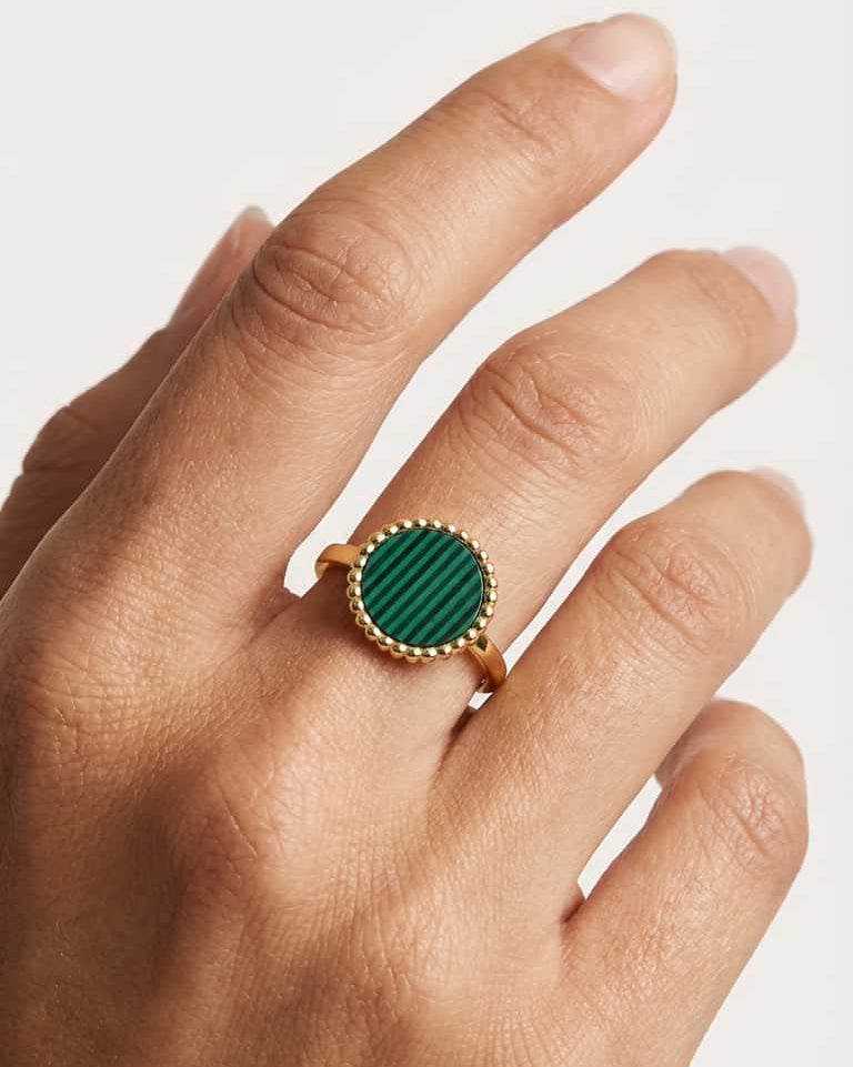 A gold beaded ring with a round green malachite stone, worn on a woman's ring finger against a clean white background.