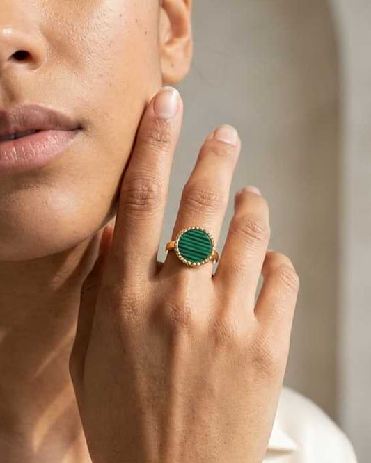 A close-up lifestyle shot of a woman wearing a gold ring with a green malachite stone on her finger, set against a soft, warm-toned neutral background.