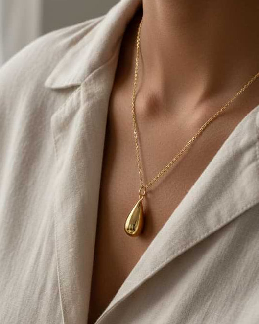 A close-up lifestyle shot of a model wearing a delicate gold chain necklace with a polished gold teardrop pendant, styled with a cream-colored linen shirt.