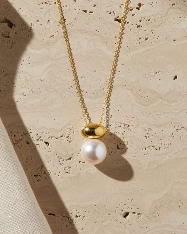 A gold chain necklace with a pendant featuring a white pearl and a polished gold bead, displayed on a textured beige stone surface with soft shadows.