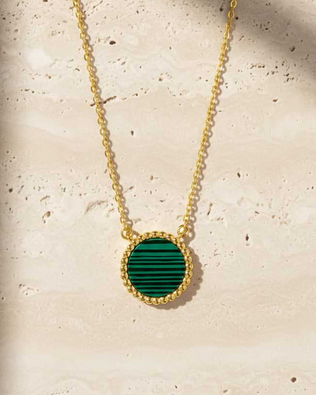 A gold chain necklace with a round pendant featuring a green malachite stone inlay and a textured gold border, displayed on a light beige stone surface.