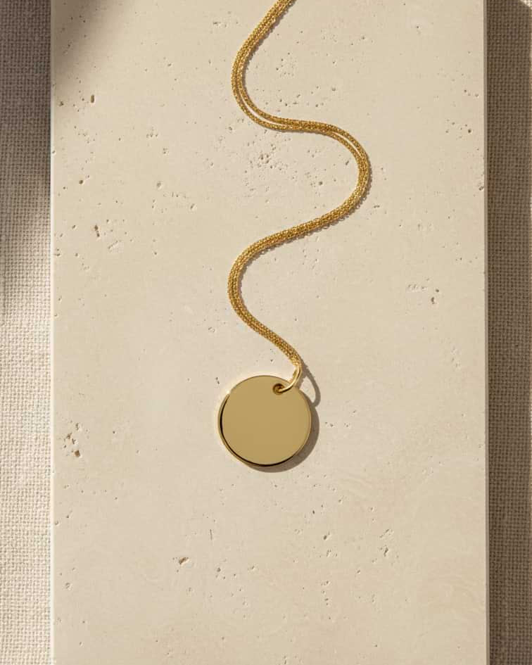 A gold chain necklace with a blank round disc pendant, displayed on a textured beige stone tile against a neutral linen background.