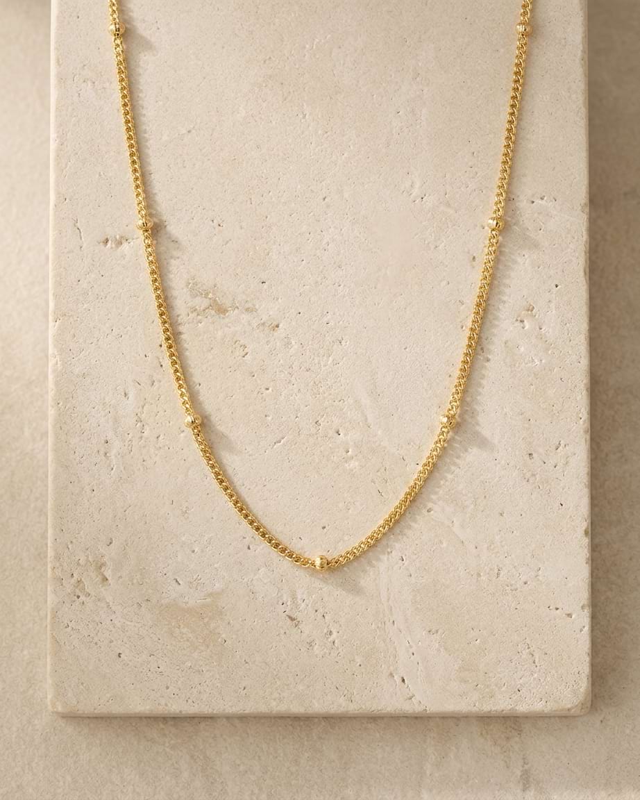 A delicate gold curb chain necklace with fine beads draped in a U-shape over a textured beige stone tile.