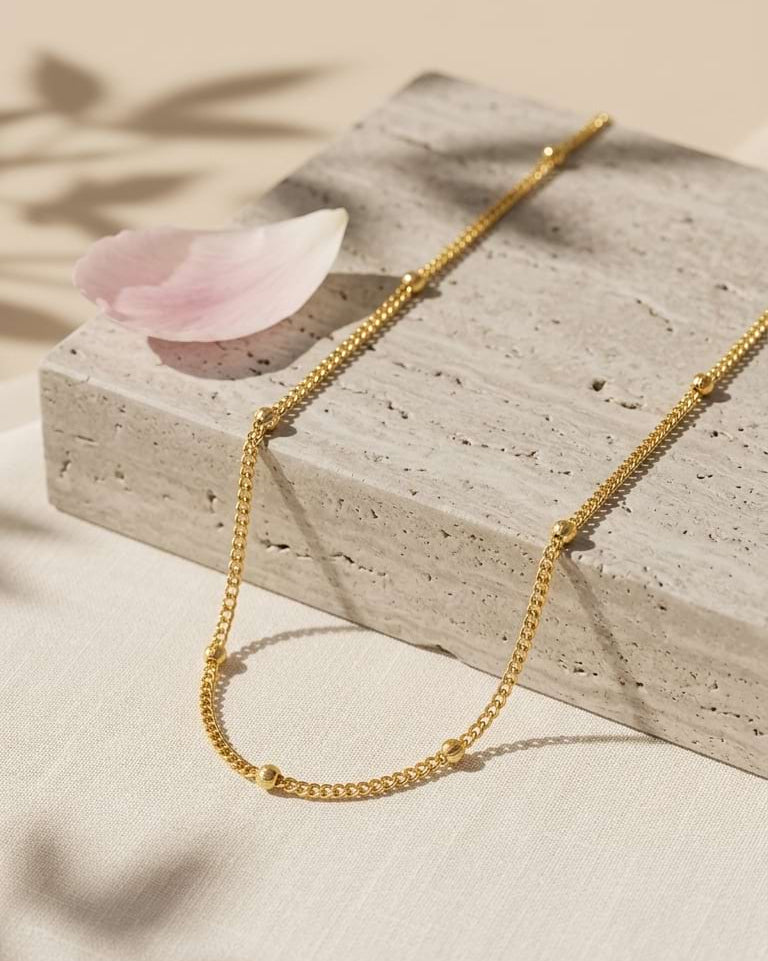 A delicate gold chain necklace draped over a textured stone slab with a soft pink petal beside it, set against a warm beige background with leaf shadows.