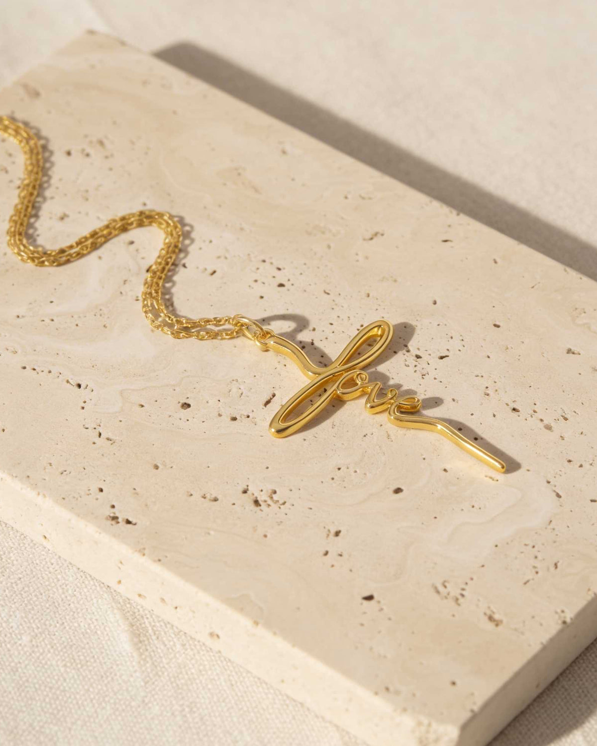 A gold chain necklace with a stylized infinity symbol pendant featuring the word 'love' in cursive, displayed on a beige stone slab.