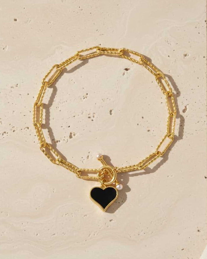 Gold bracelet with a black heart charm on a beige surface