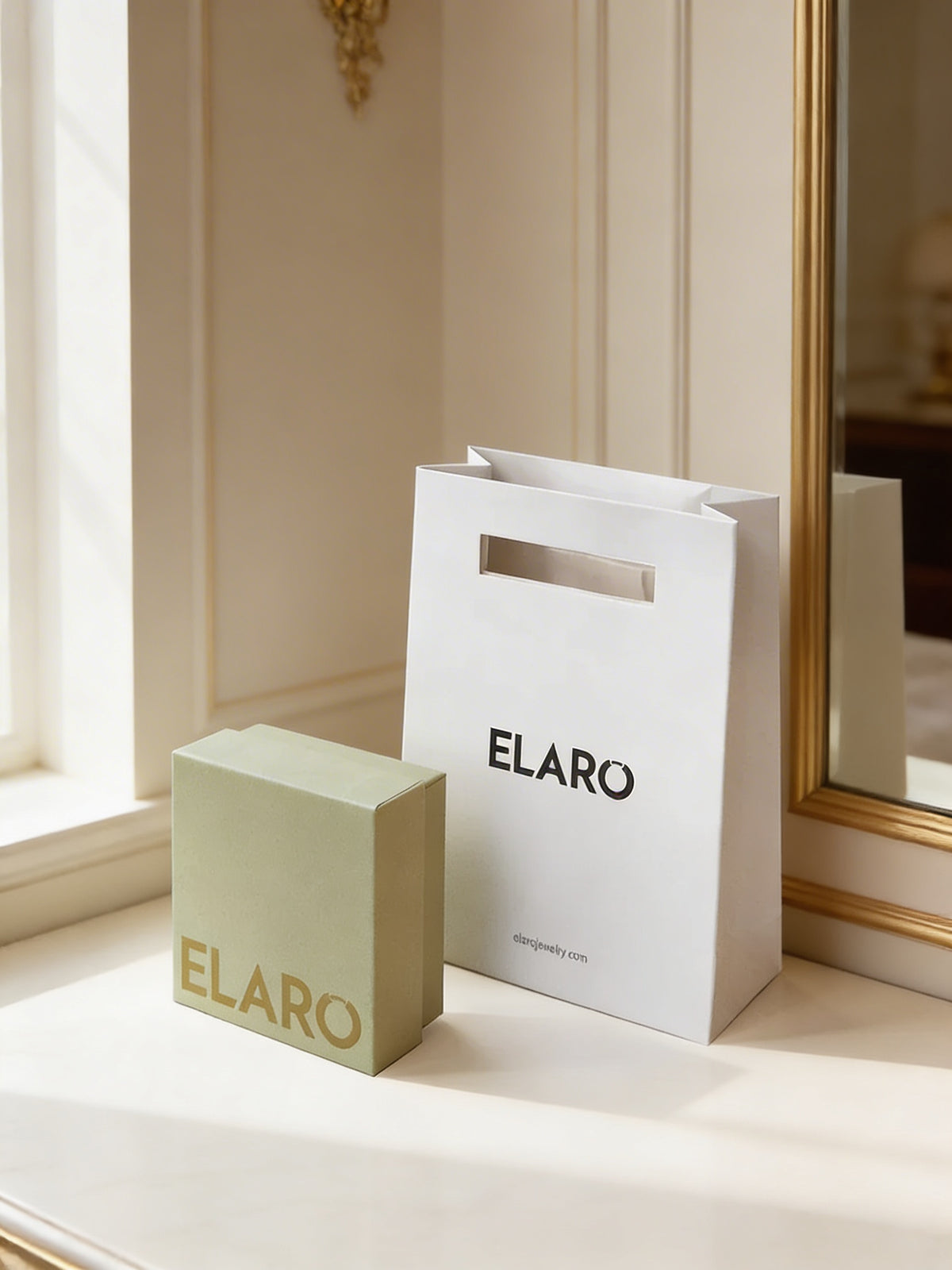 A white ELARO paper shopping bag and a light sage green rectangular box with gold ELARO lettering, placed on a cream-colored windowsill in a softly lit interior setting.