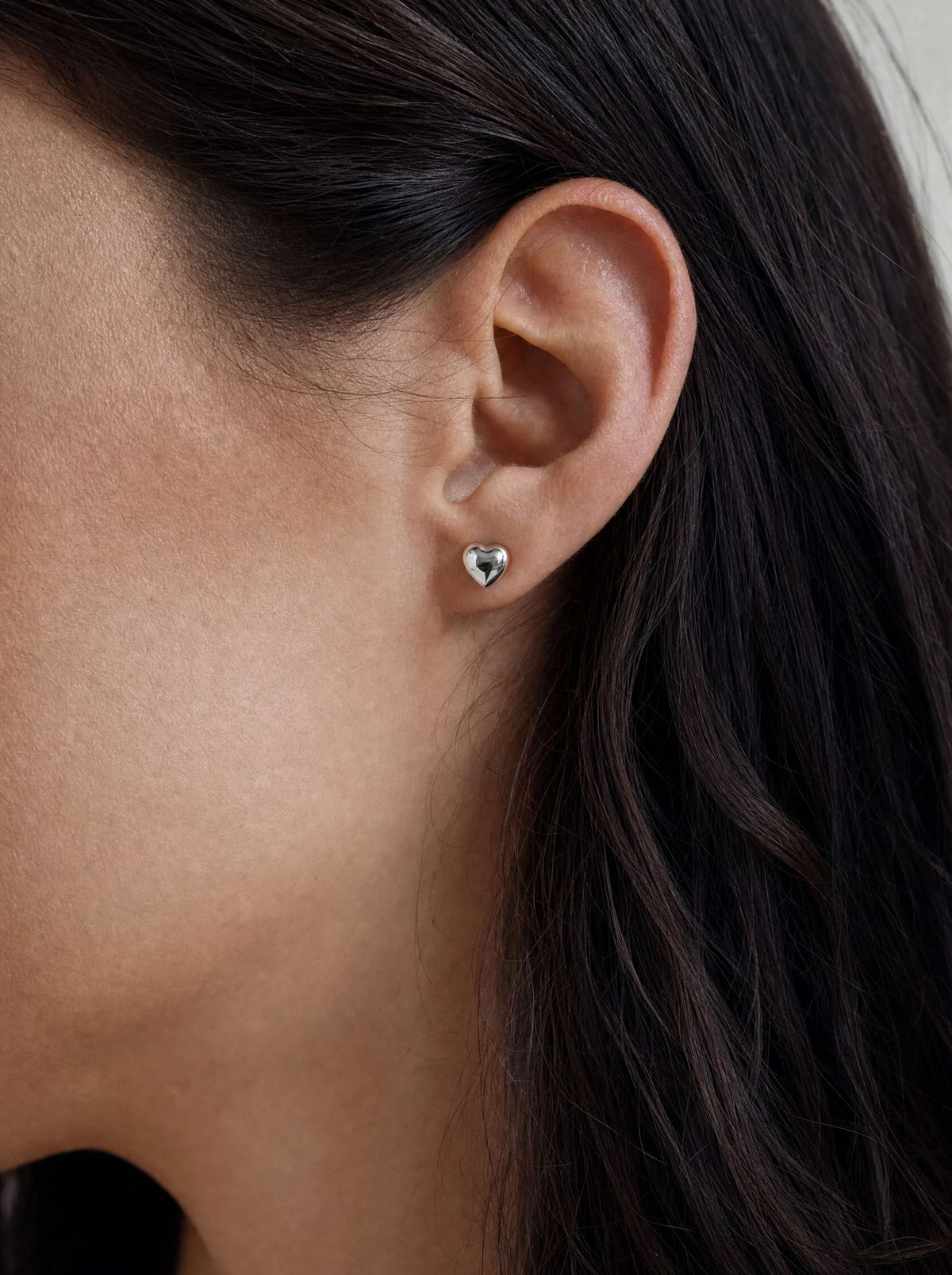 A close-up of a model's ear wearing a small white gold heart-shaped stud earring.